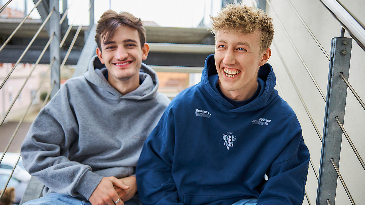 2 junge Männer in Hoodies sitzen lachend auf einer Treppe