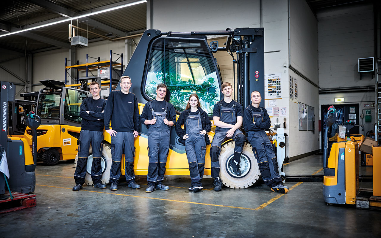 Gruppenbild von Azubis in Arbeitsanzügen vor einem großem, gelben Gabelstapler