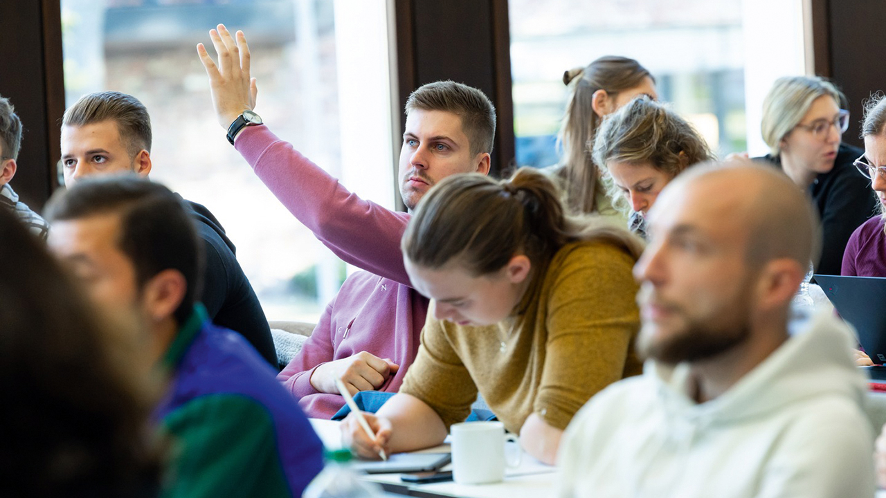 Studierende in einer Lehrveranstaltung. Eine Person schreibt mit, eine meldet sich, eine weitere schaut interessiert. 