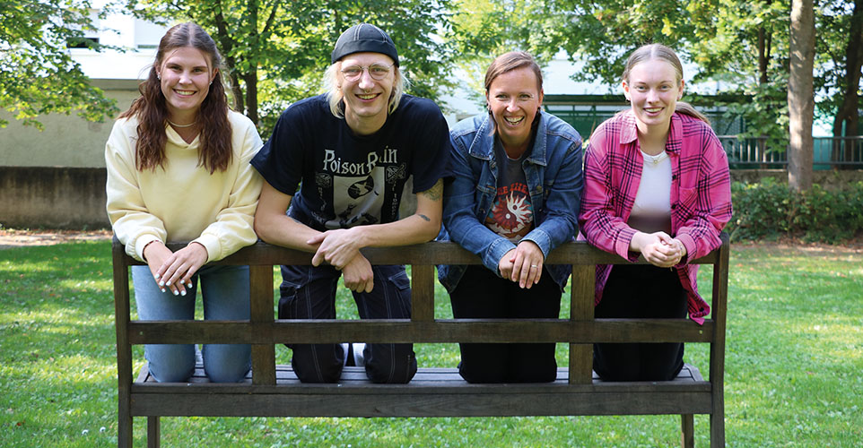 Vier Personen knien auf der Sitzfläche einer Bank und lächeln in die Kamera.