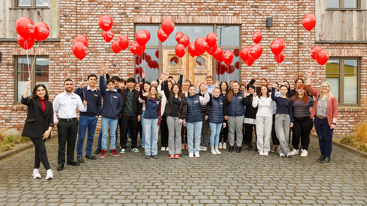 Gruppenfoto mit vielen Menschen. Sie stehen vor einer Klinkerfassade und halten alle rote Luftballons in die Höhe