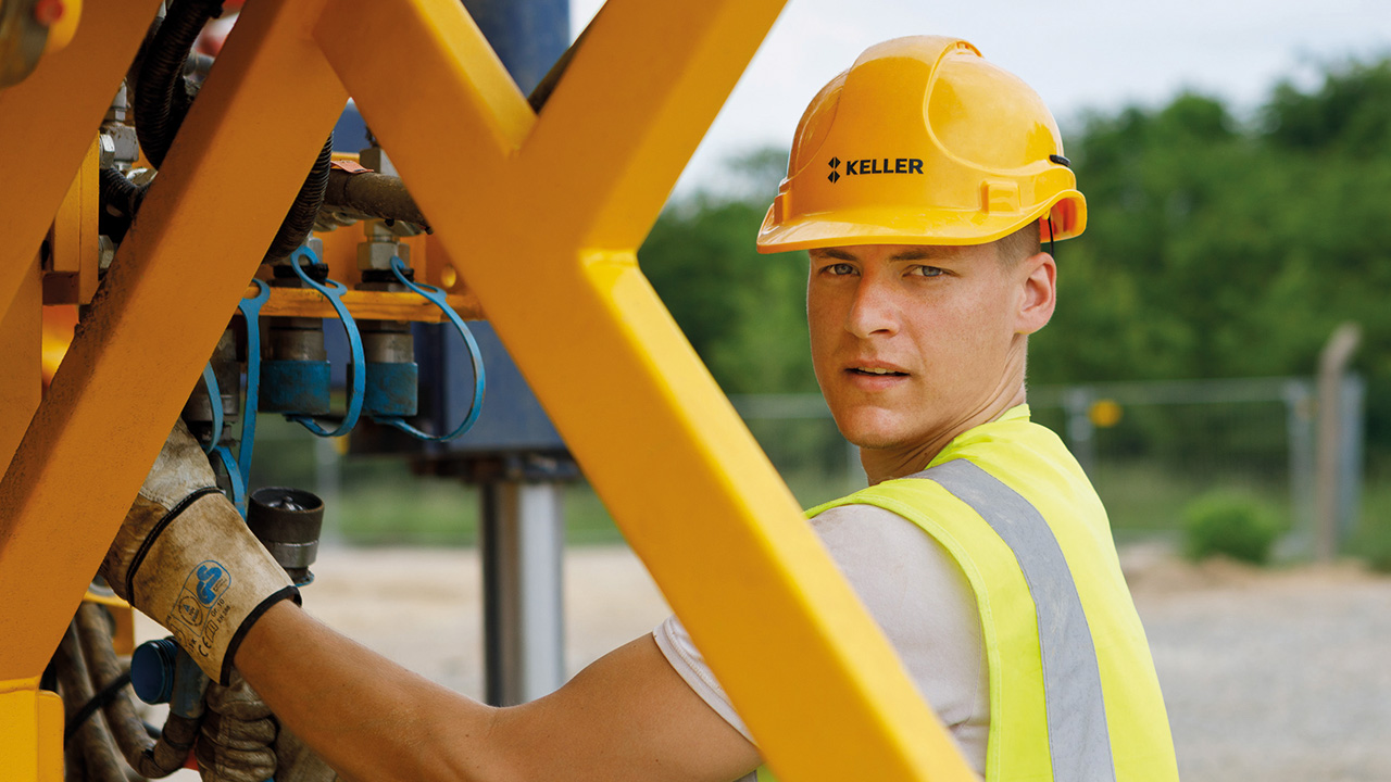 Junger Mann mit Warnweste und Bauhelm, der die Aufschrift KELLER trägt, guckt in die Kamera