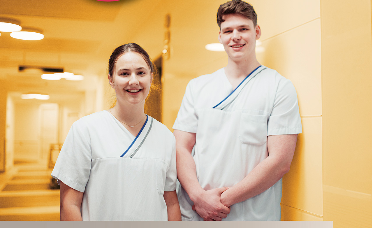 Zwei junge Menschen in Pflege-Kleidung, die in einem Krankenhausflur stehen und in die Kamera lächeln