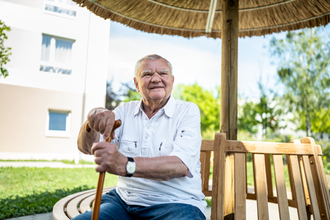 Älterer Mensch mit Stock in der Hand sitzt auf einer Bank unter einem Sonnenschirm