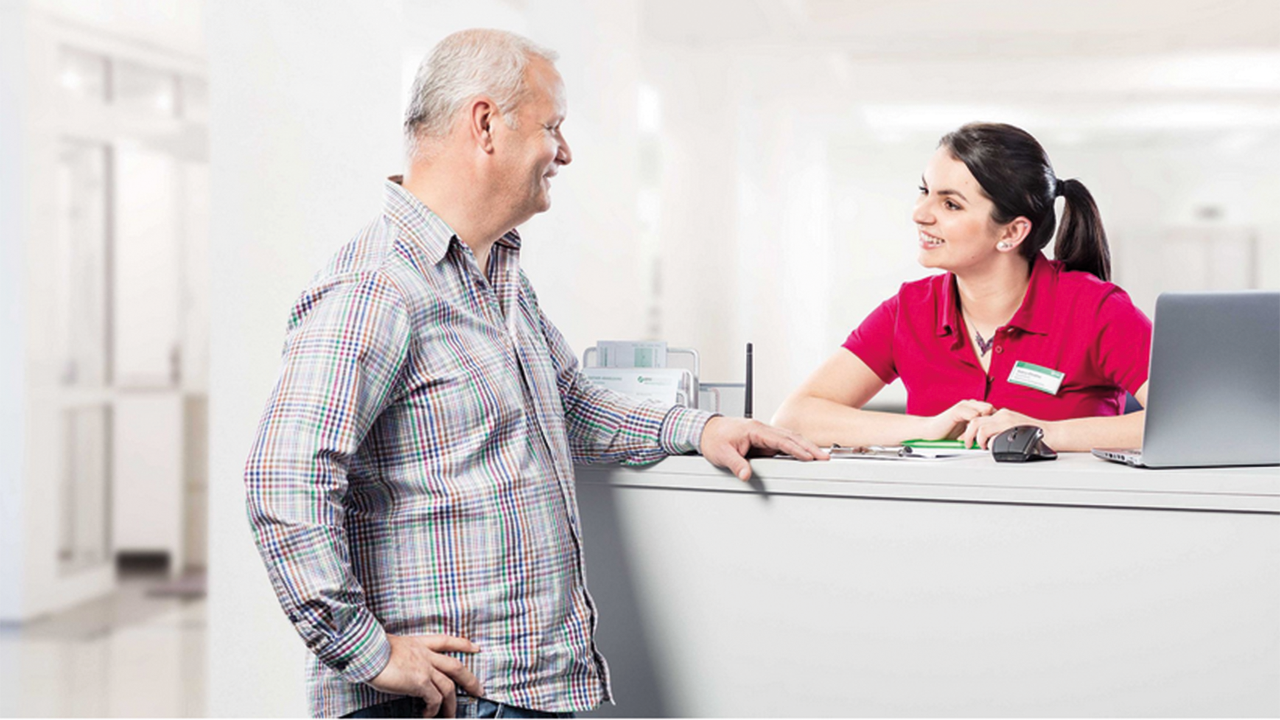 Älterer Mann steht an der Anmeldung einer Arztpraxis. Eine junge Mitarbeiterin lächelt ihn freundlich an.