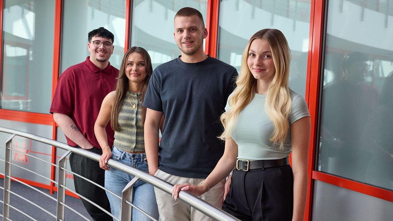 Gruppe von vier Personen steht an einem Geländer vor Fenstern mit roten Rahmen