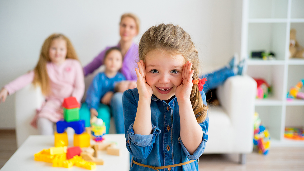 Das Bild zeigt ein Kind im Vordergrund was die Hände an das Gesicht hält. Im Hintergrund sitzen zwei weitere Kinder zusammen mit einer erwachsenen Person auf einer hellen Couch. Ein weiteres Kind liegt auf dem Schoß der erwachsenen Person. 