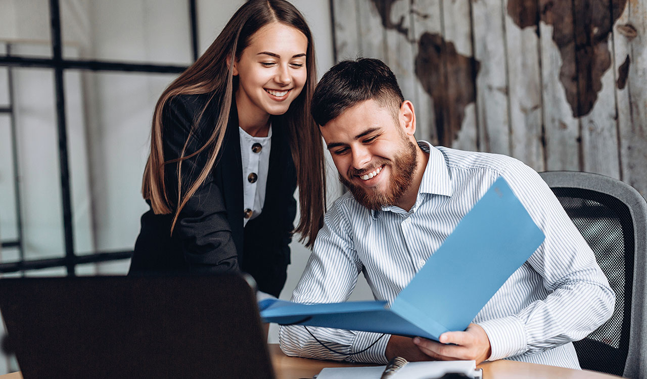 Zwei Personen an einem Schreibtisch. Eine Person steht leicht nach vorne gebeugt, die andere sitzt und hält einen blauen Ordner. Beide lächeln.