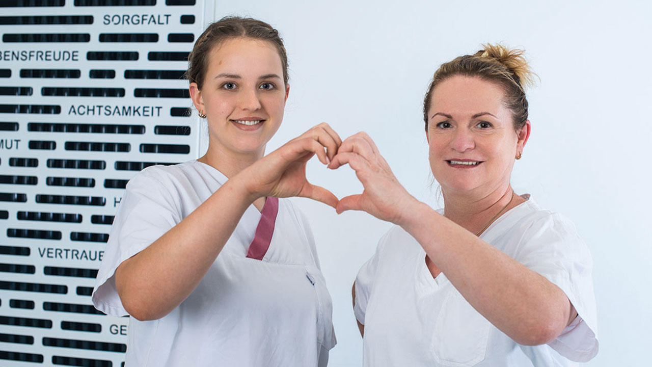 Zwei lächelnde Personen in weißer Pflegebekleidung formen mit ihren Händen ein Herz