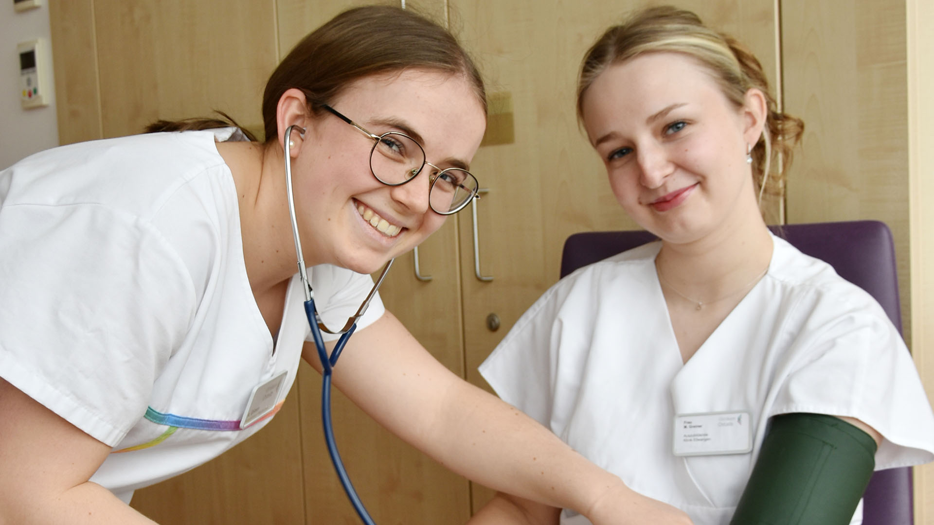Eine Person misst einer anderen Person den Blutdruck. Beide lächeln in die Kamera.