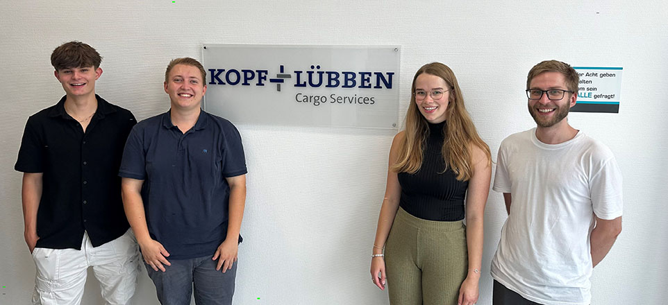 Vier junge lächelnde Personen vor einem Schild mit dem Firmenlogo