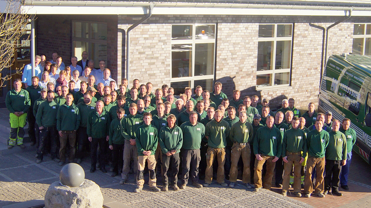 Viele Personen stehen in Arbeitskleidung der Krebs & Suhr GmbH & Co. KG vor einem Gebäude und blicken hoch in die Kamera. 
