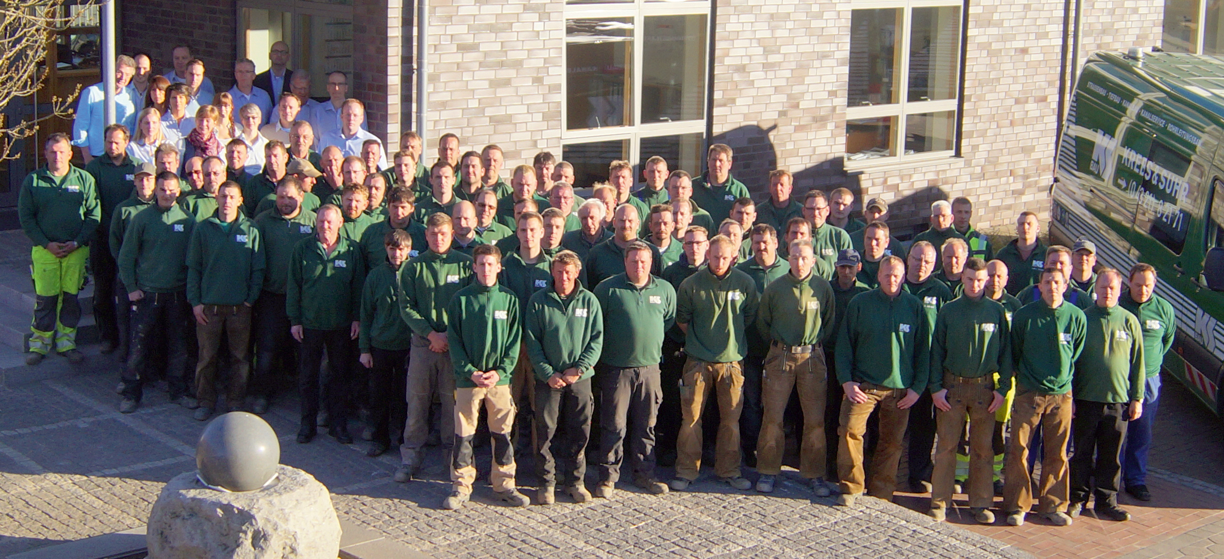 Viele Personen stehen in Arbeitskleidung der Krebs & Suhr GmbH & Co. KG vor einem Gebäude und blicken hoch in die Kamera. 