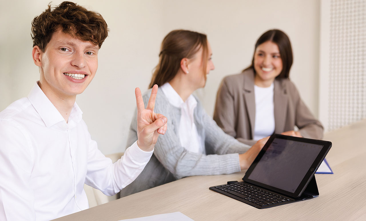 Drei Jugendliche sitzen an einem Tisch, ein Junge in weißem Hemd zeigt das Victory-Zeichen.