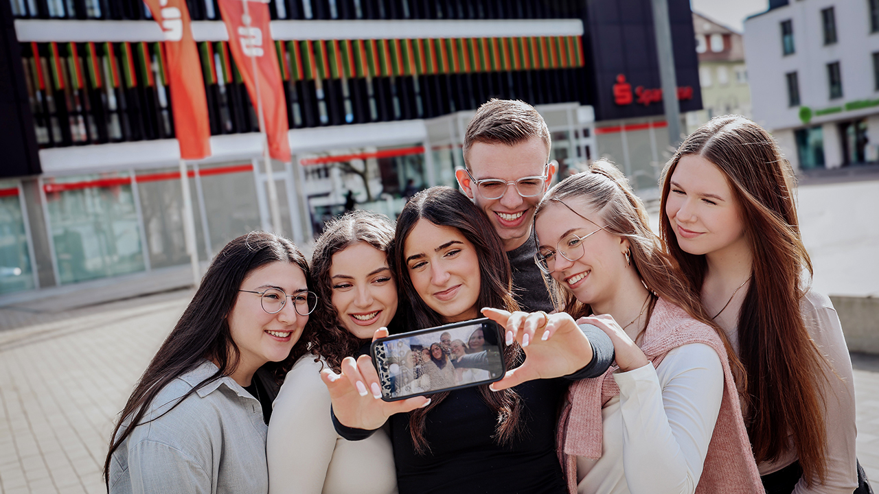 Das Bild zeigt eine Personengruppe die vor einer Sparkassenfilialie ein Selfie macht. 