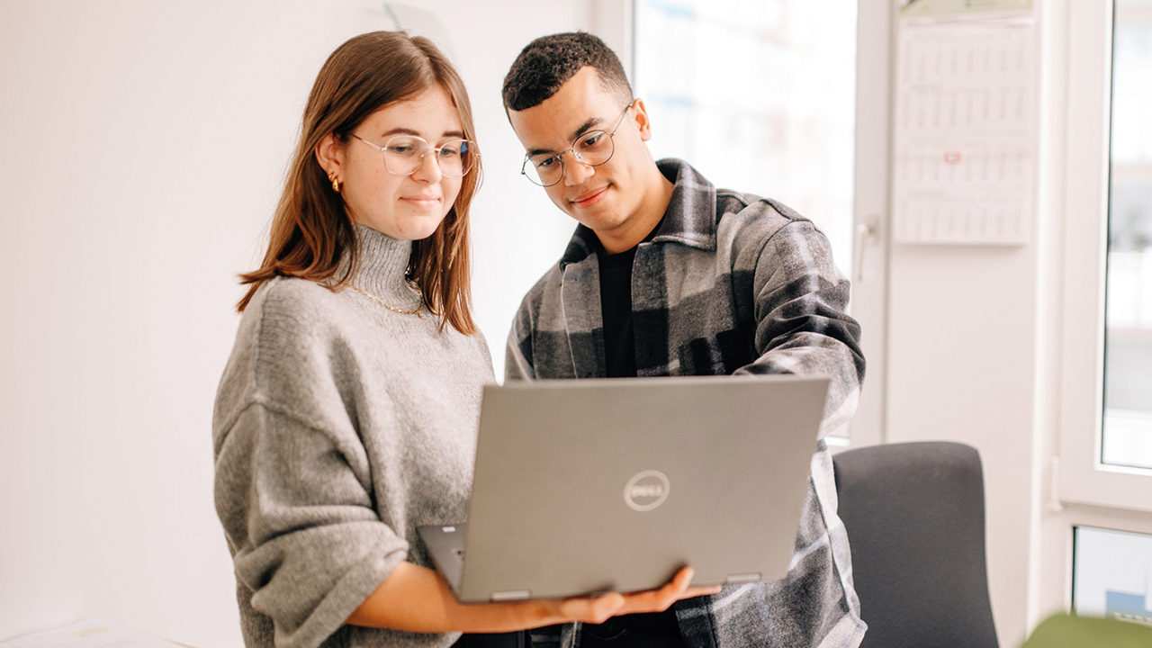 Eine junge Frau steht in einem Büro und hat einen aufgeklappten Laptop auf dem Unterarm. Sie schaut auf den Bildschirm. Neben ihr steht ein junger Mann und zeigt ihr etwas auf dem Laptop. 