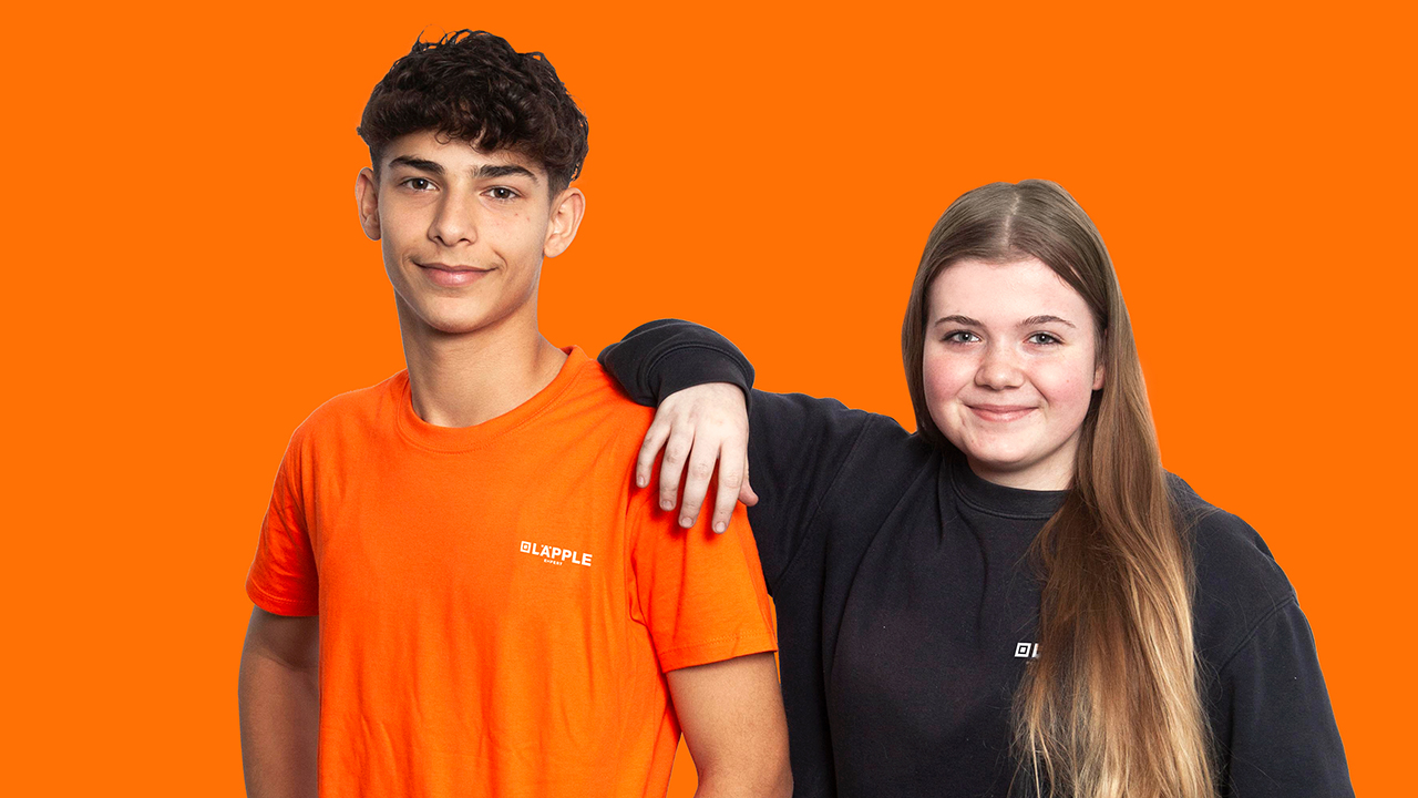 Zwei Personen vor einem orangen Hintergrund gucken in die Kamera und lächeln. 