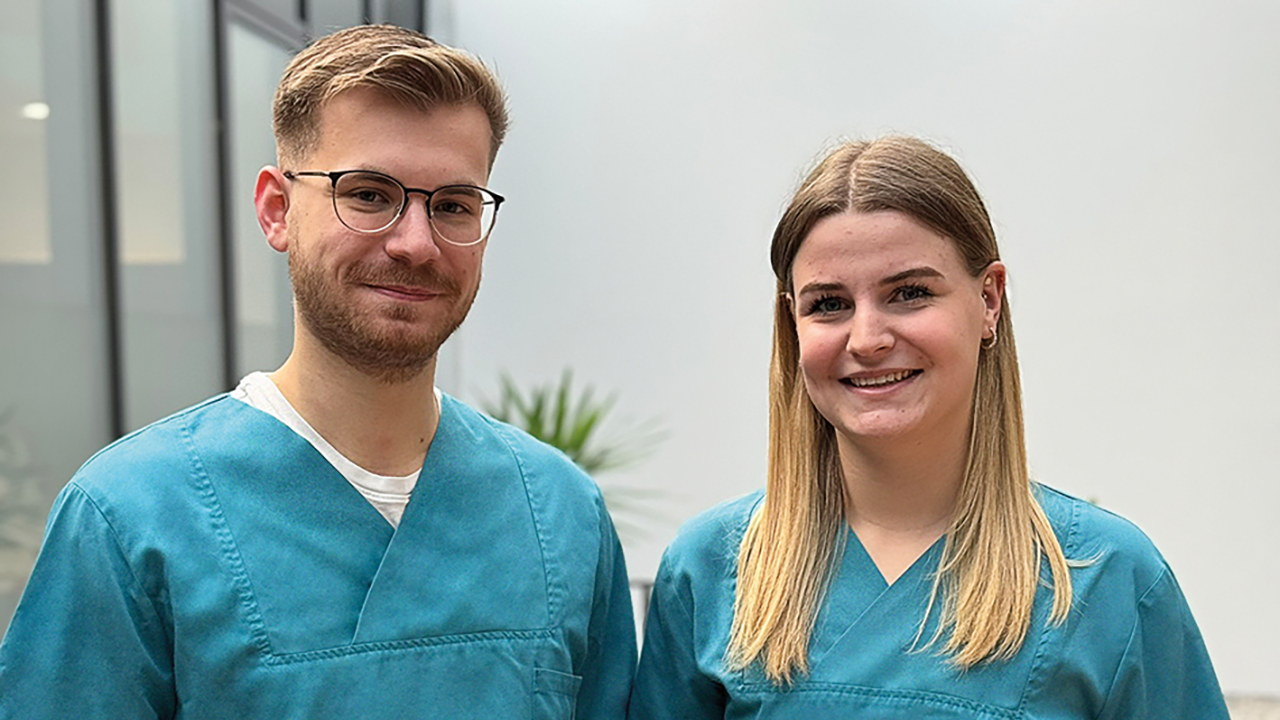 Zwei Personen tragen medizinische Arbeitskleidung und lächeln in die Kamera. 