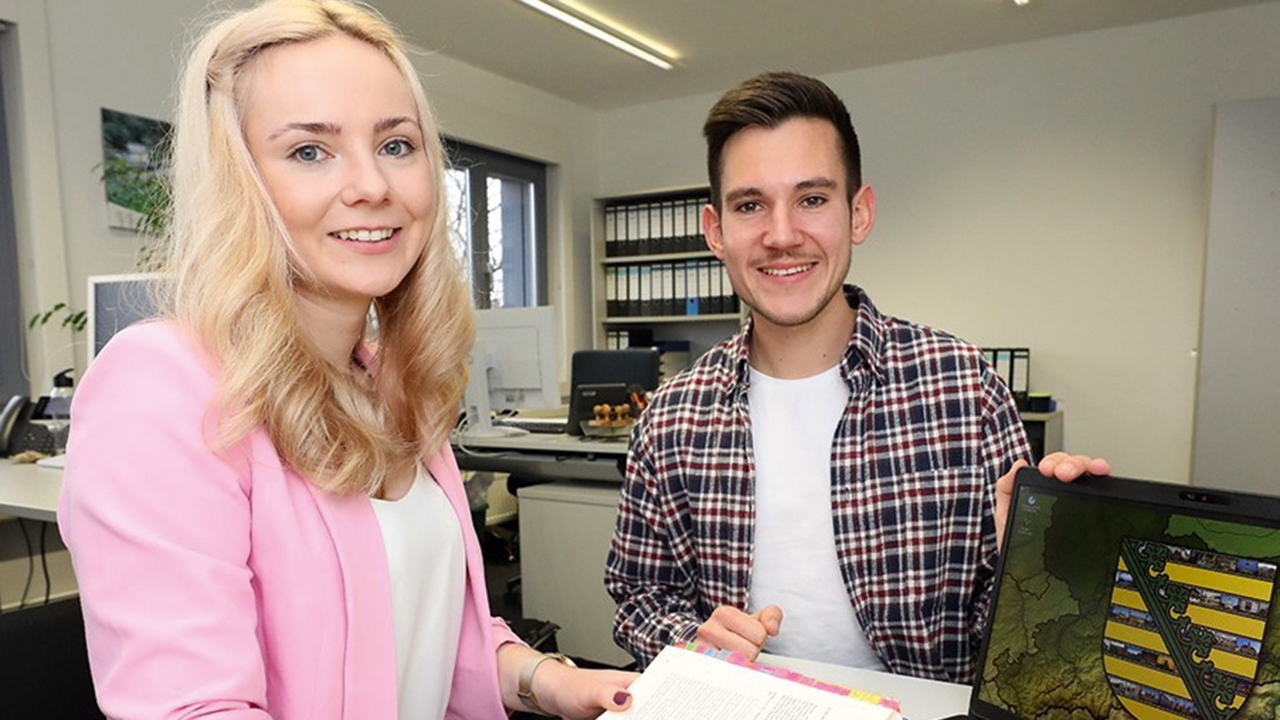 Bild einer jungen Frau und eines jungen Mannes im Büro. Sie hat ein Buch aufgeschlagen vor sich, er präsentiert einen aufgeklappten Laptop auf dem das Logo des Freistaates Sachsen zu sehen ist.