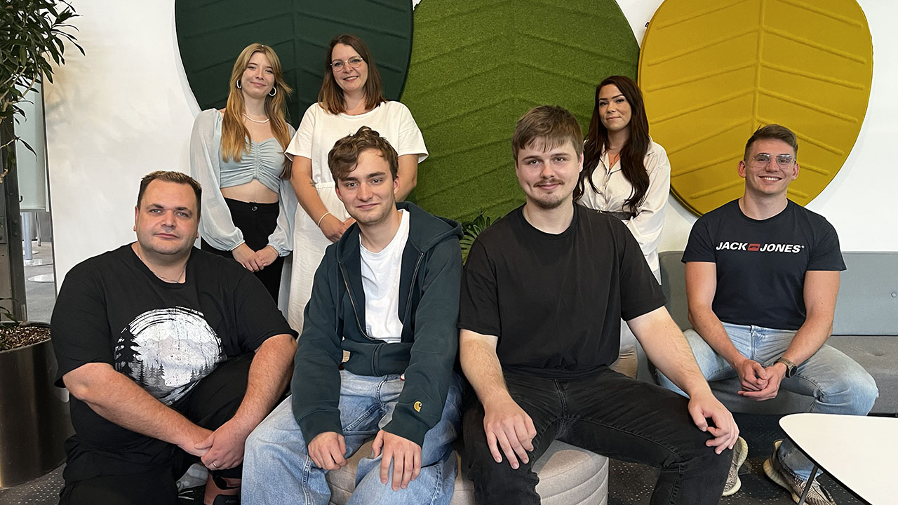 Im Vordergrund sitzen vier junge Personen, im Hintergrund stehen drei junge Personen, sie lächeln in die Kamera