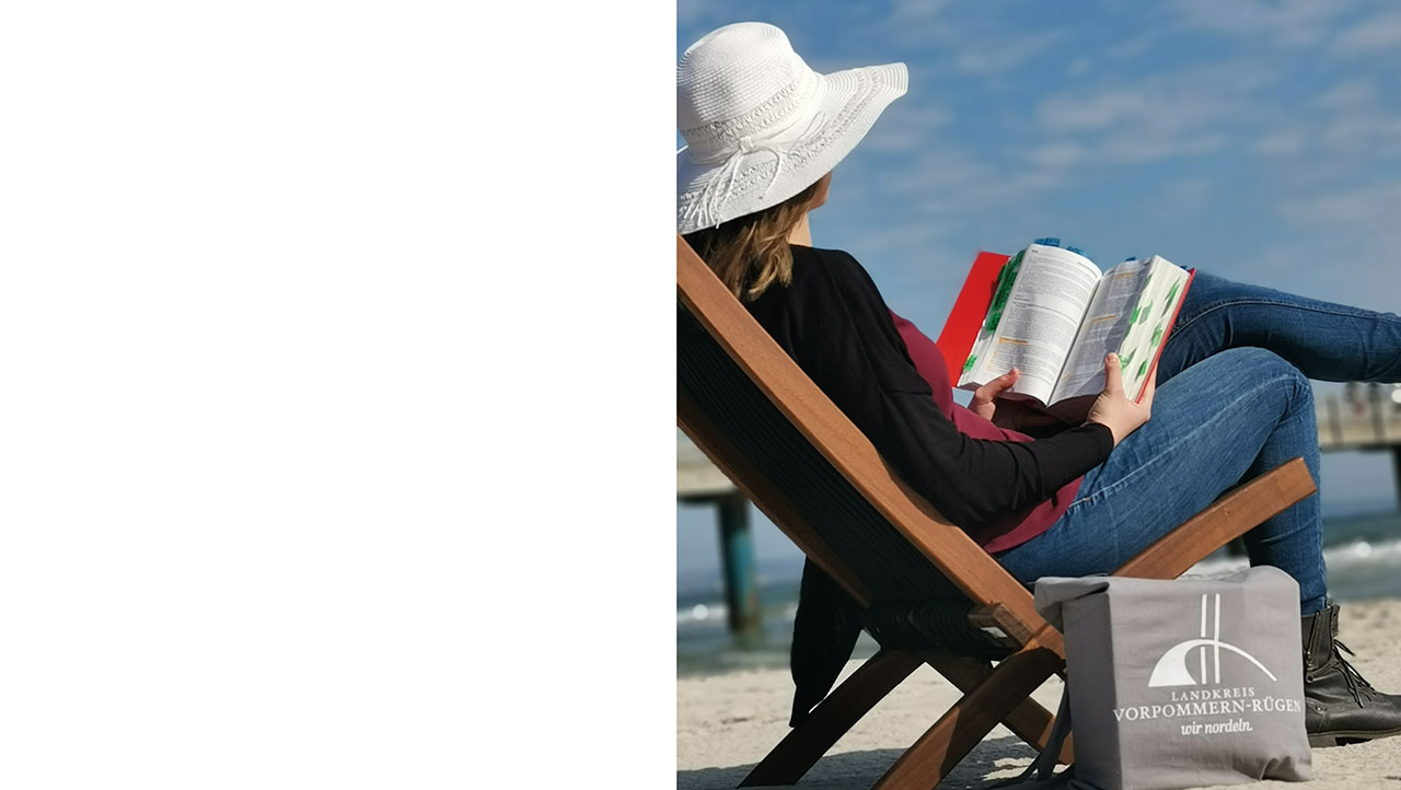 Person mit Hut und Buch in der Hand im Liegestuhl am Strand