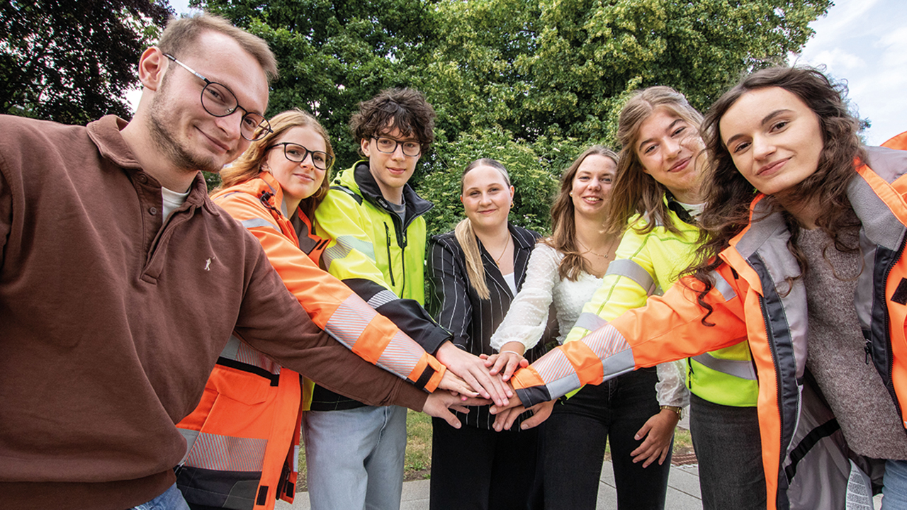 7 junge Menschen stehen nebeneinander im Halbkreis und strecken jeweils eine Hand aus, so dass alle Hände übereinander liegen. 