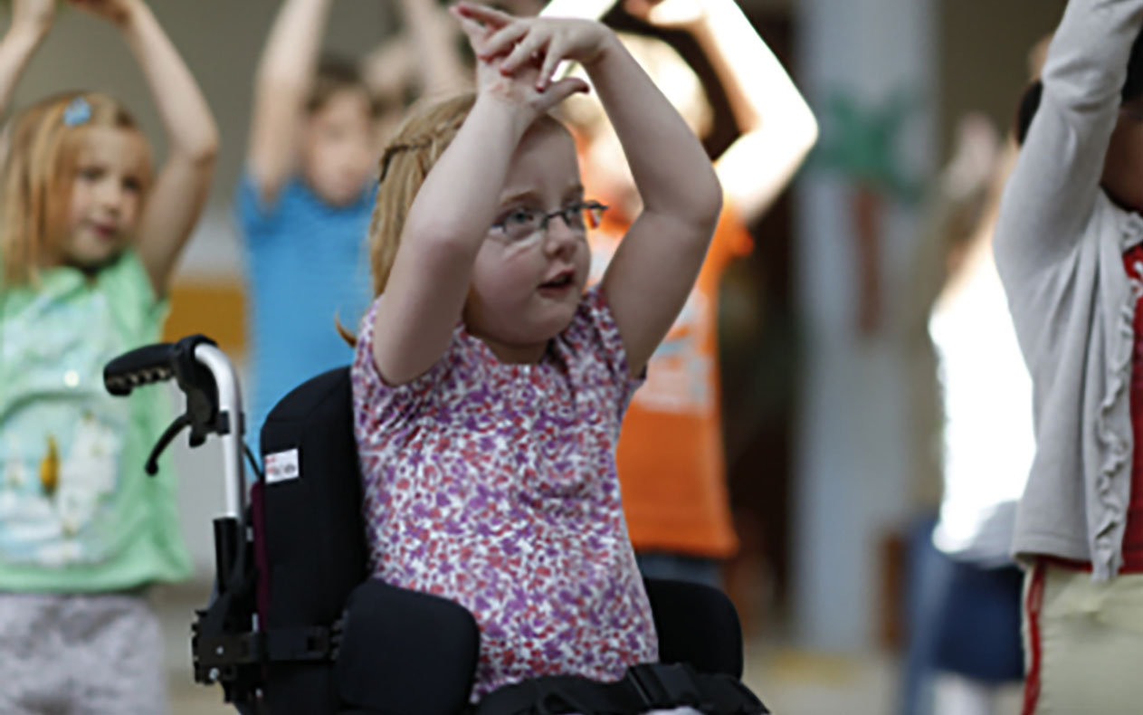 Kleines Mädchen mit roten haaren und Brille sitzt im Rollstuhl und hebt die Hände über den Kopf.