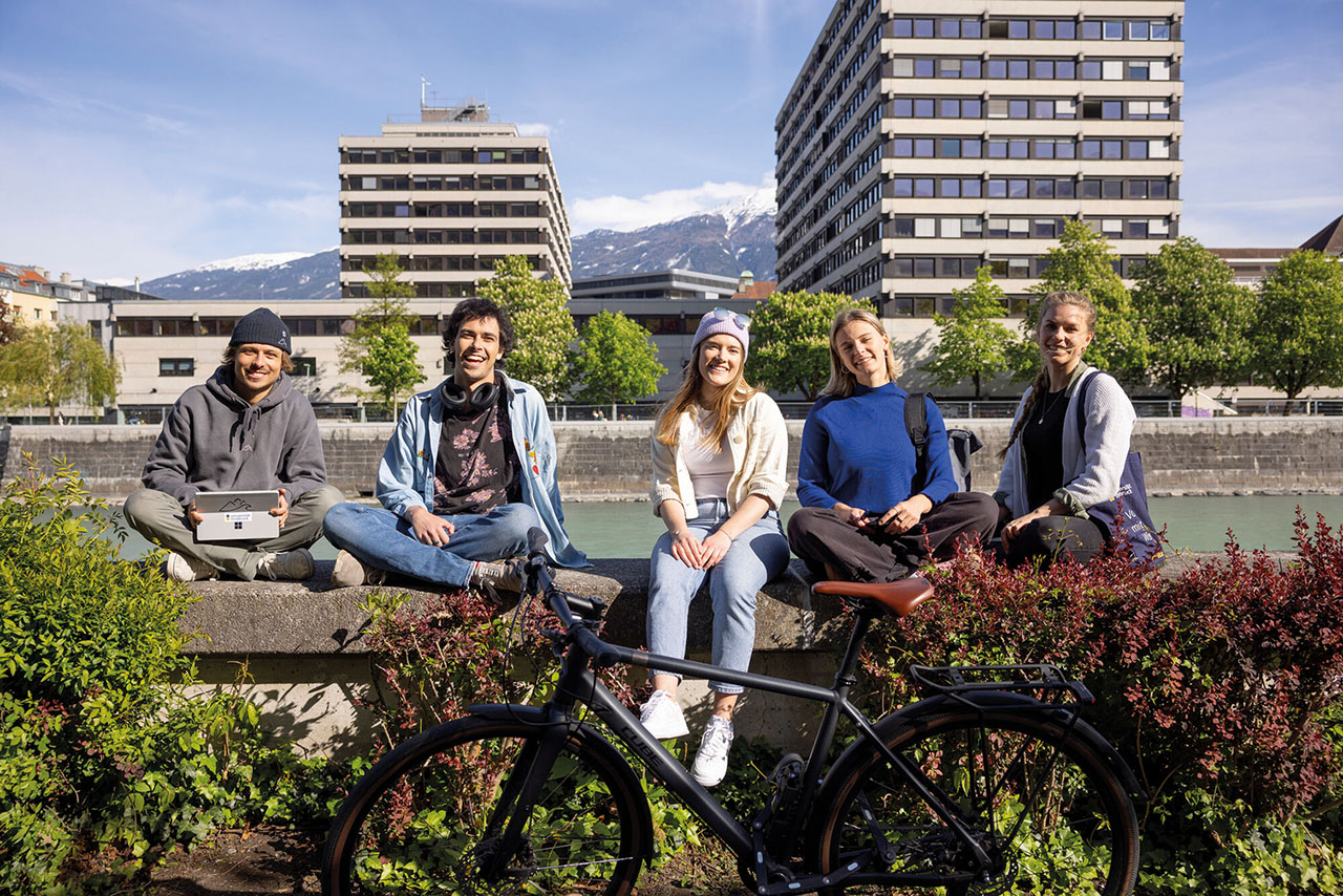 Fünf Personen sitzen lächelnd auf einer Mauer am Wasser, umgeben von Pflanzen. Im Hintergrund sind hohe Gebäude und Berge zu sehen.