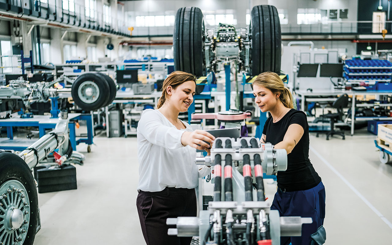 Zwei Personen arbeiten gemeinsam an einer großen Maschine in einer hellen Werkhalle mit weiteren technischen Geräten und Flugzeugrädern im Hintergrund.