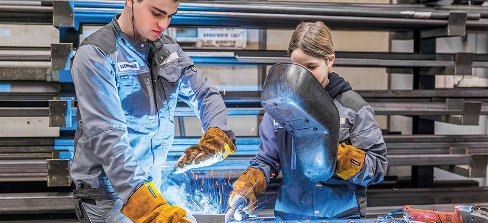 Zwei Personen in Schutzkleidung schweißen. Die rechte Person hält eine Schweißerbrille, die linke schützt die Augen mit Hilfe der Hände.