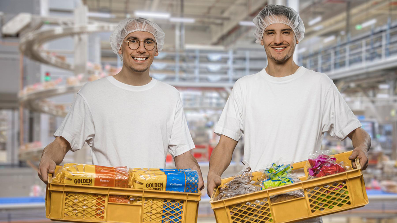 Zwei Mitarbeiter halten Stiegen mit Brotprodukten in den Händen. Sie tragen Produktionskleidung.