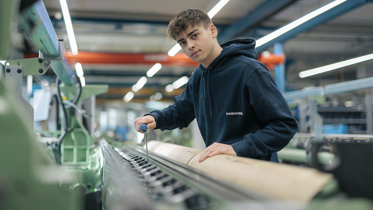 Eine junge Person steht in einer Werkhalle an einer großen Maschine. Die Person trägt einen dunklen Pullover mit dem Schriftzug „DORNIER“ und arbeitet mit einem Schraubendreher an der Maschine.