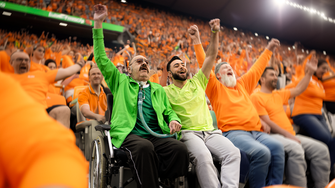 Zwei Menschen mit grünen Shirts sitzen in einem Stsadion umgeben von Menschen in orangenen Shirts