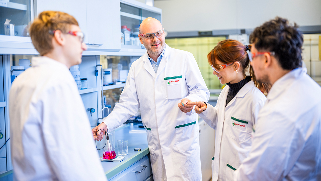 Szene im Labor. Ausbilder erklärt 3 dabei stehenden Azubis etwas. Alle in weißer Schutzkleidung.