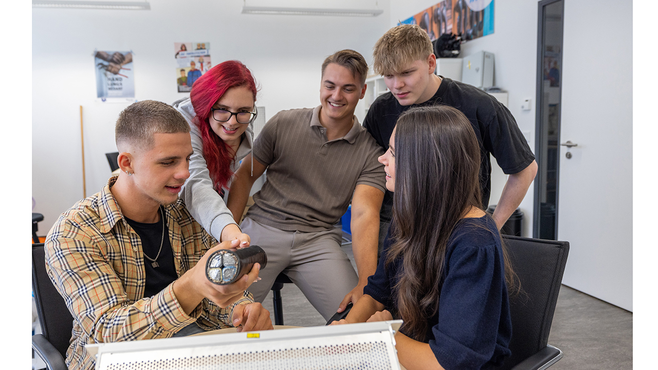 Fünf junge Menschen schauen sich ein dickes Stück Kabel an und sitzen dabei an hinter einem Schreibtisch