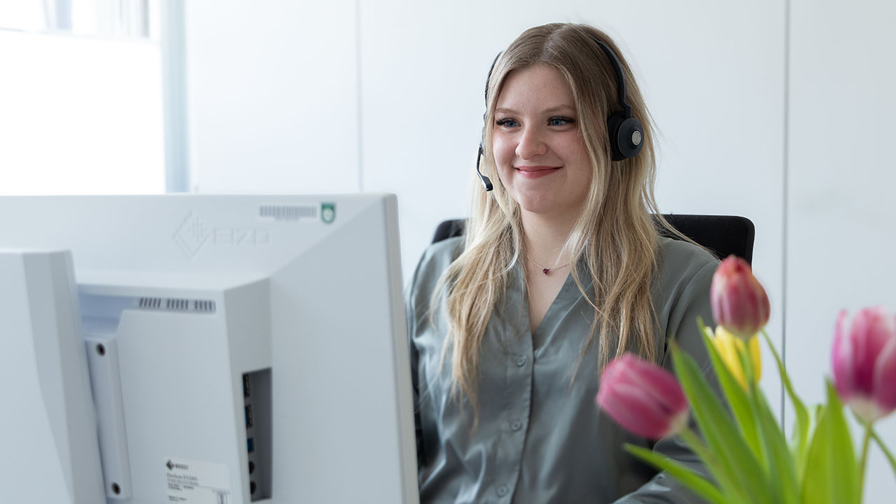 Person sitzt lächelnd hinter einen Computer, rechts im Vordergrund sind drei Tulpen.