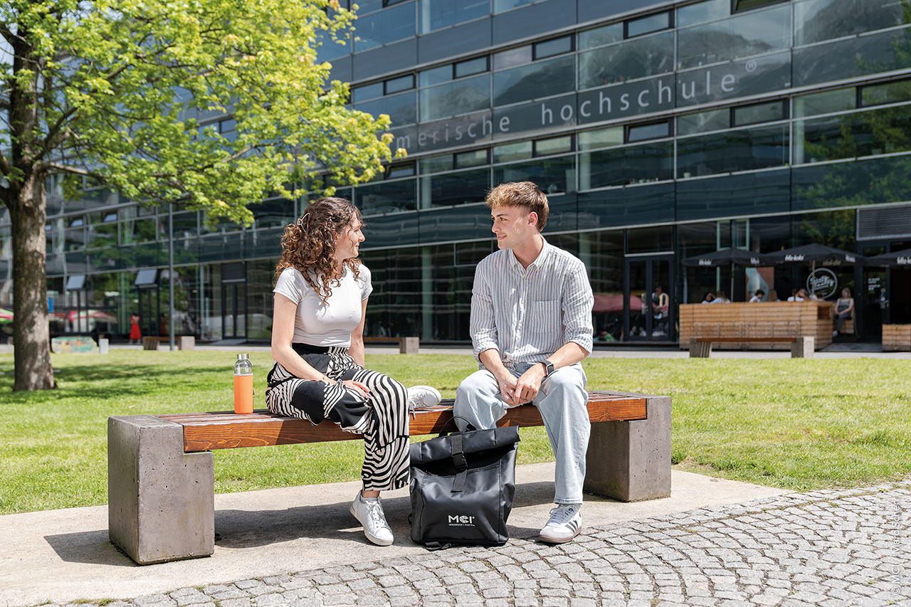 Zwei Personen sitzen auf einer Bank vor einem modernen Gebäude mit Glasfassade.