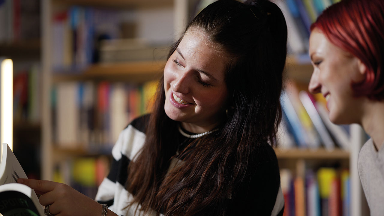 Zwei junge Personen schauen vor einer Bücherwand zusammen in ein Buch