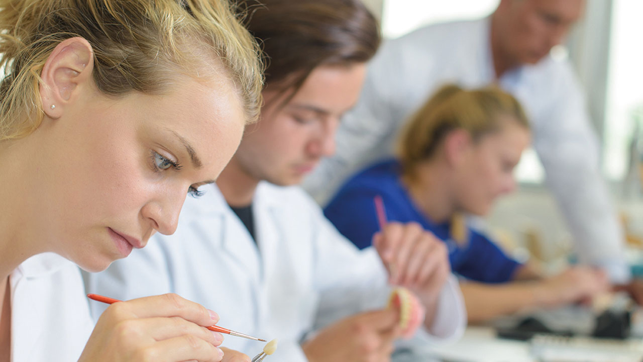 Junge Person arbeitet mit einem Pinsel an einem Zahnersatz, im Hintergrund zwei weitere Personen bei der Arbeit an Prothesen