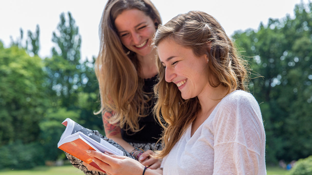 Zwei junge lächelnde Menschen mit langen Haaren sitzen im Grünen und blättern in einem Buch