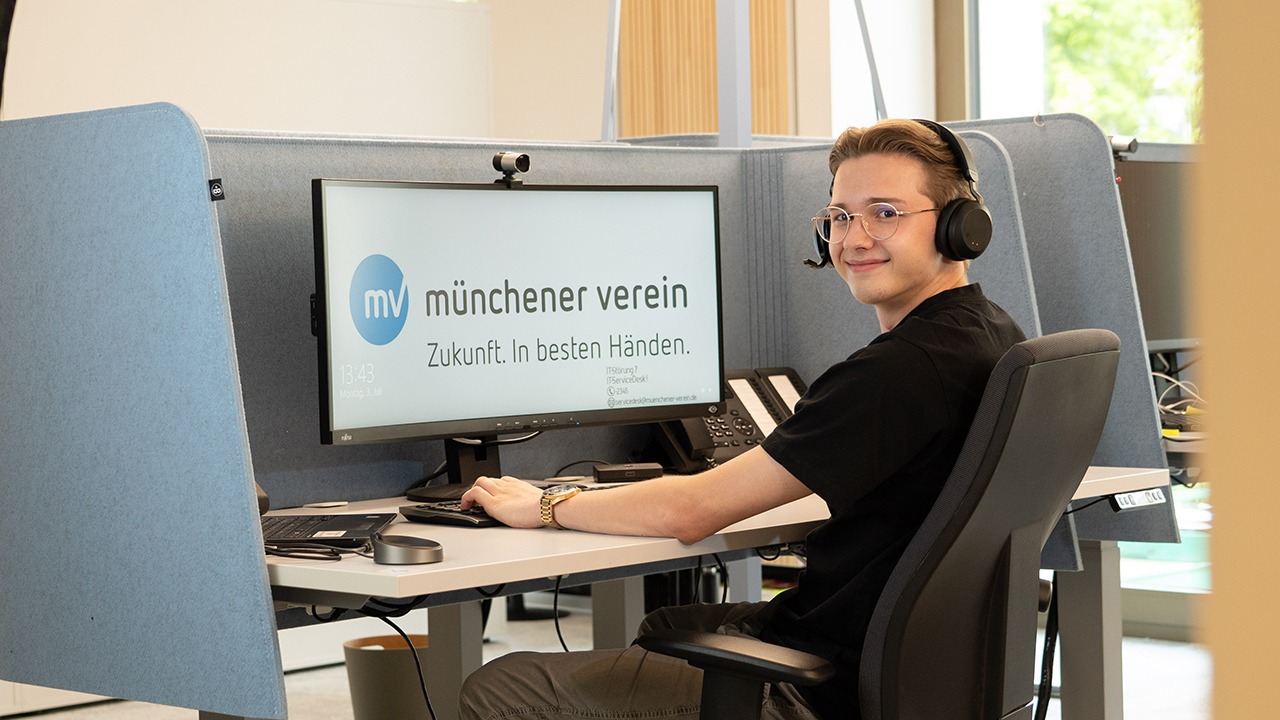 Junger Mensch sitzt mit Headset an einem Schreibtisch. Vor ihm ein Bildschirm mit dem Logo "Münchner Verein"