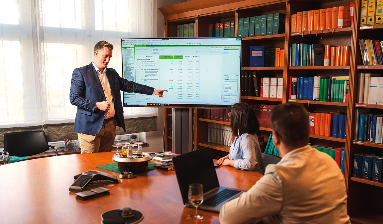 Bürositutation. Eine person steht an einem Flatscrren und erklärt zwei anderen sitzenden Personen eine Tabelle.