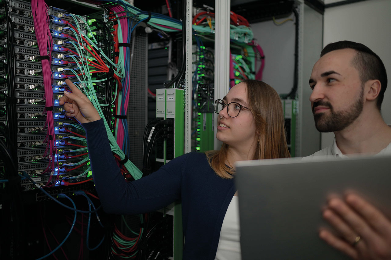 Das Bild zeigt zwei Personen in einem Serverraum. Eine Person zeigt auf Kabel und Geräte in einem Serverschrank, die andere hält einen Laptop. Überall sind bunte Netzwerkkabel und leuchtende Lichter zu sehen.
