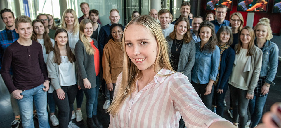 In der Mitte eine Person die in die Kamera lächelt im Hintergrund eine große Personengruppe. 