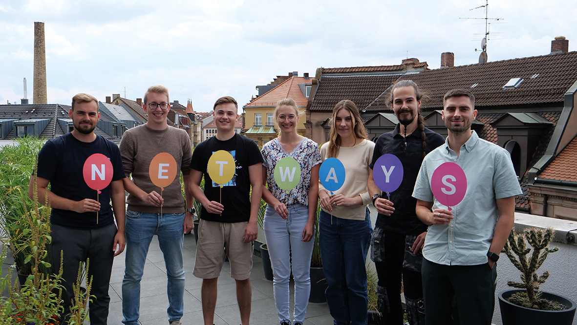 Gruppenbild auf einer Dachterrasse. Jede und jeder hält ein bundes Schild mit je einem Buchstaben des Wortes Netways in der Hand.