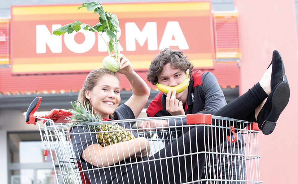 Eine junge Frau sitzt in einem Einkaufswagen, hat eine Ananas im Arm und hält sich einen Kohlrabi auf den Kopf. Hinter dem Wagen steht ein junger Mann, der sich eine Banane vor den Mund hält. Im Hintergrund der Eingang eines Norma Marktes.