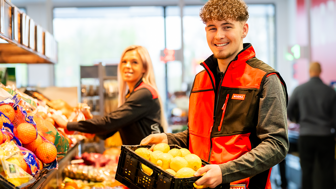 Obstabteilung, zwei junge Mitarbeitende sortieren frisches Obst in die Auslage, beide lächeln freundlich den Betrachter an.