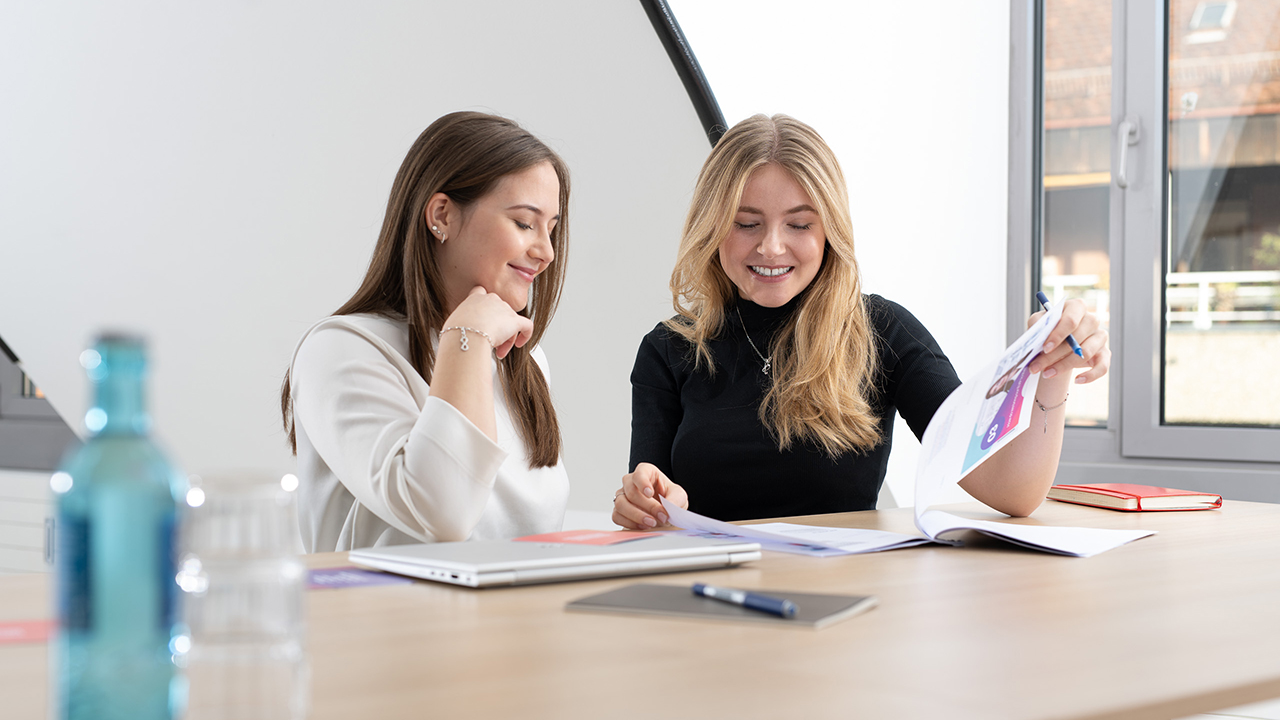 Zwei junge Mitarbeiterinnen an einem Schreibtisch, bei der Arbeit im Büro.