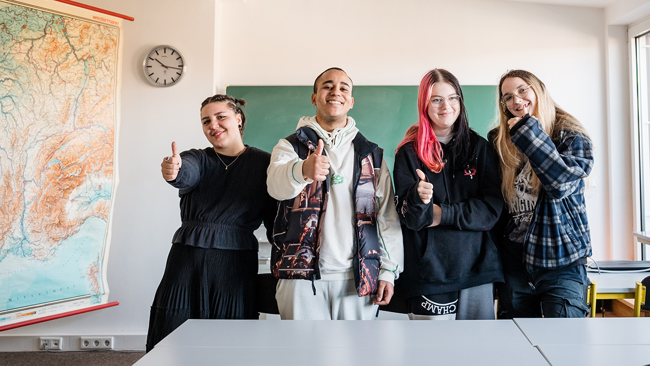 4 Schüler/innen stehen in einem Klassenzimmer vor einer Tafel und zeigen den Daumen nach oben. Links im Bild sieht man eine Landkarte. An der Wand hängt eine Uhr.