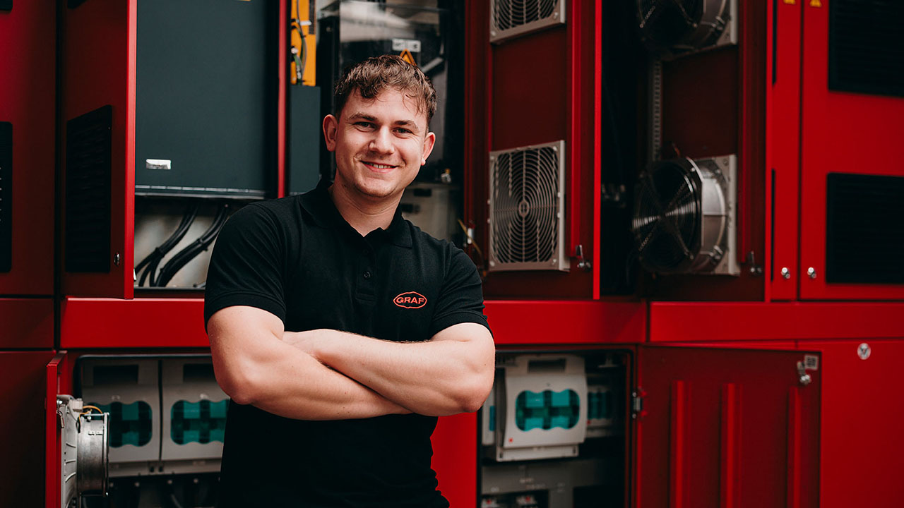 Junger Mensch im schwarzen Polsohirt mit Firmenlogo steht vor einer elektronischen Anlage und lächelt in die Kamera